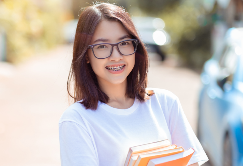 Smiling young woman with braces and glasses holding books outdoors