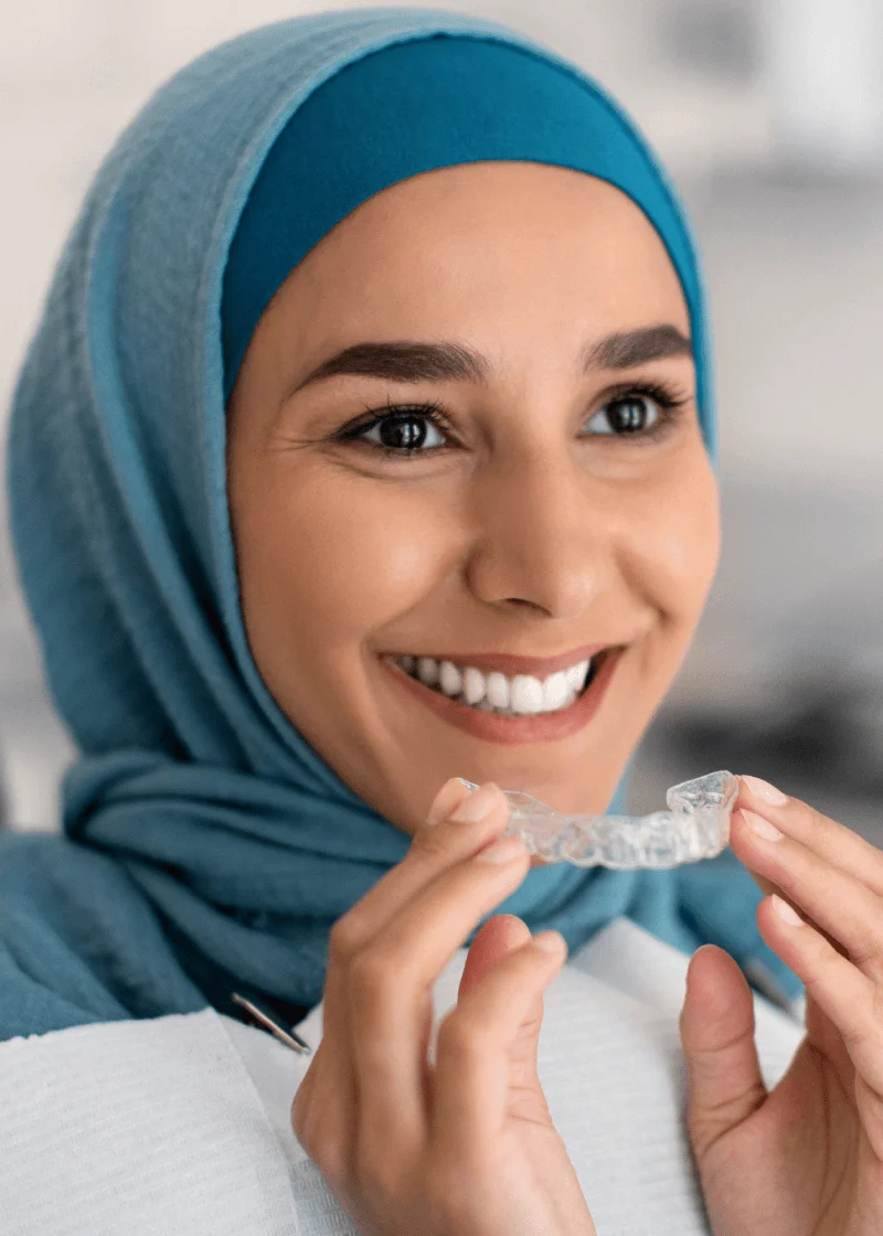 Smiling woman in a teal hijab holding a clear Invisalign aligner tray