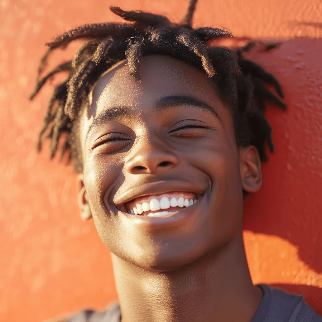 Young man with locs laughing with eyes closed against an orange wall