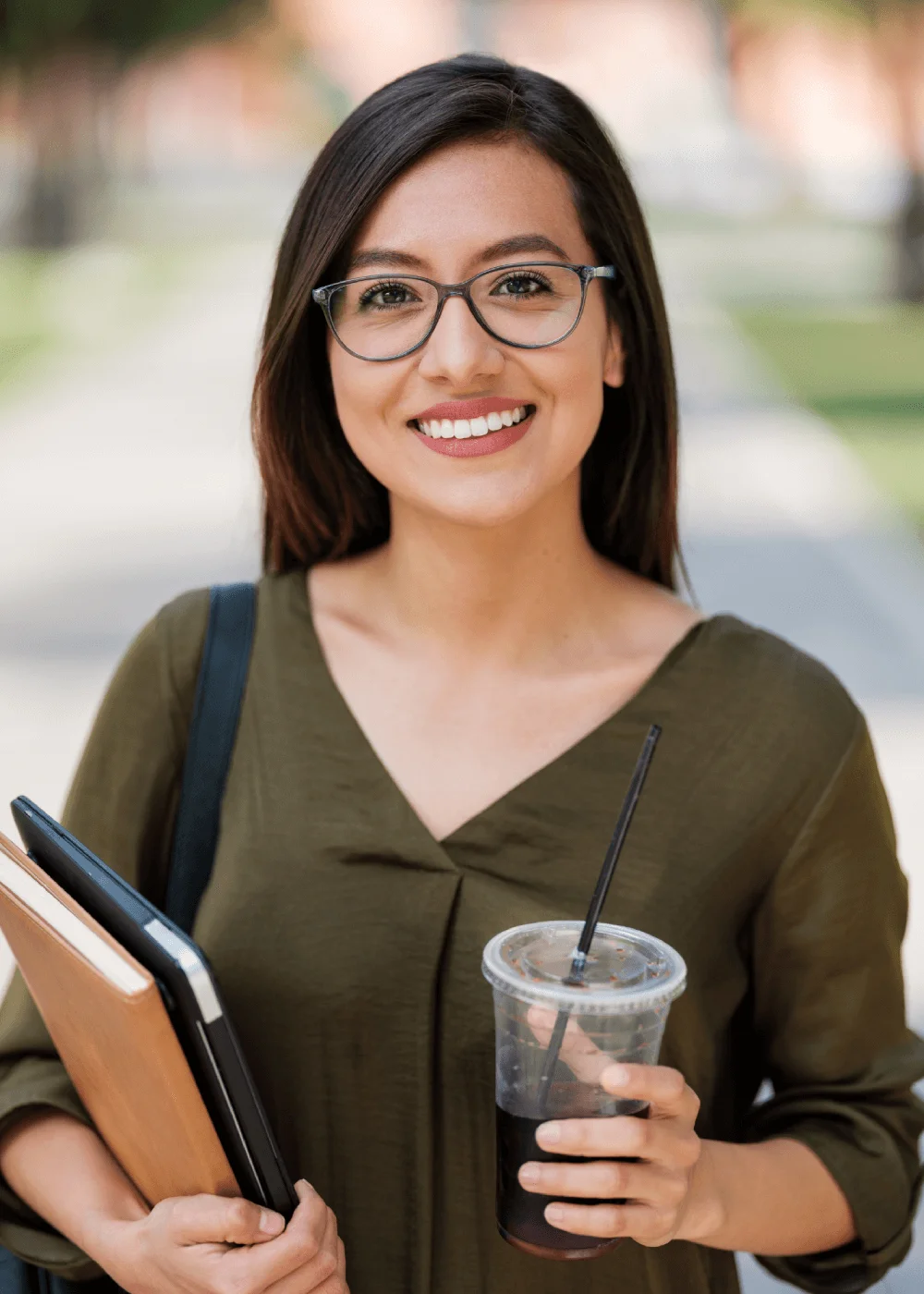 Smiling young woman in glasses holding books and an iced coffee outdoors