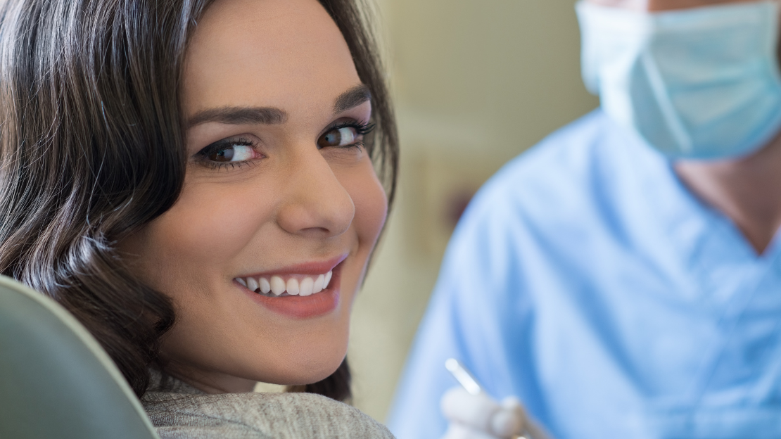 Smiling woman looking over her shoulder from a chair, a person in a mask and blue scrubs behind her